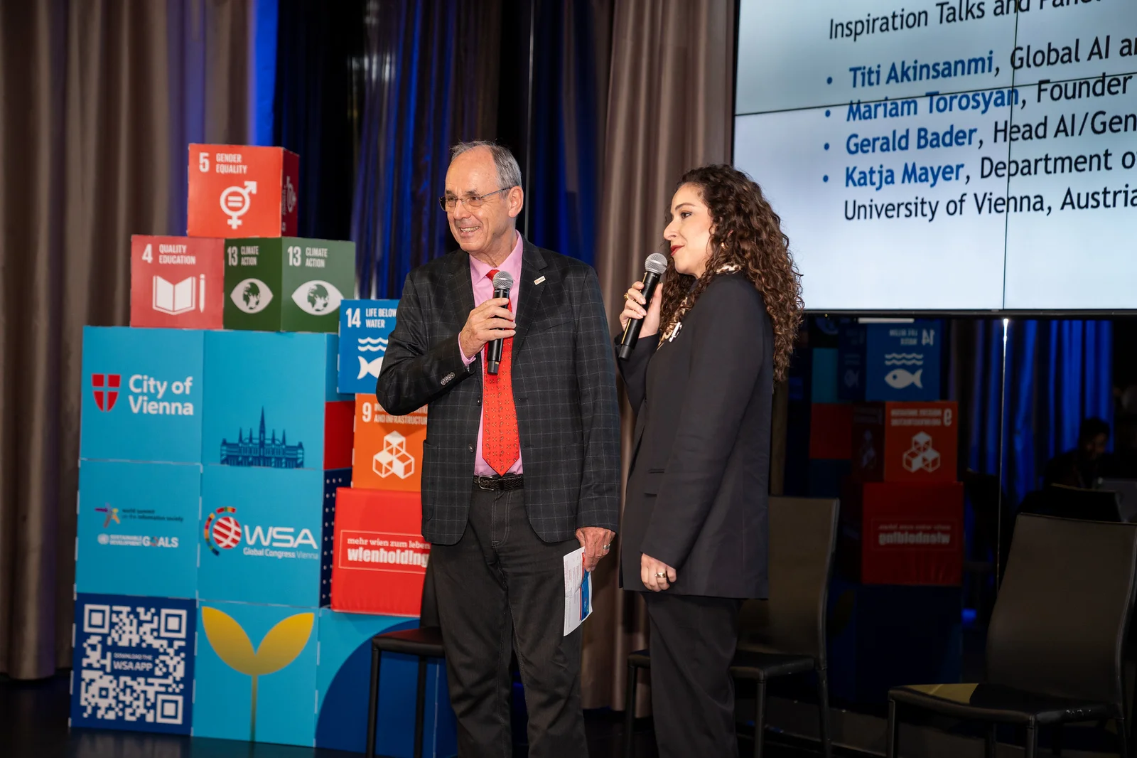 Two presenters holding microphones beside the Sustainable Development Goals display in Vienna.